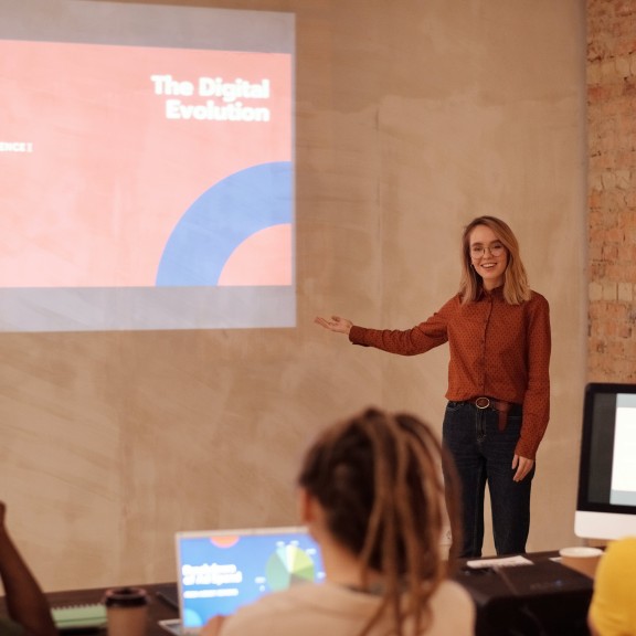 A woman in a long-sleeved shirt and pants is presenting to three students on "The Digital Evolution". The presentation is projected onto the wall, one student its in front of a PC, one has a laptop open, and one has a notebook. The room is tan with a brick wall next to it.