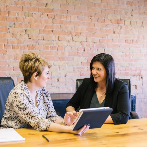 two businesswomen looking at an ipad sitting in office chairs and an office wooden meeting table with a brick wall behind them.