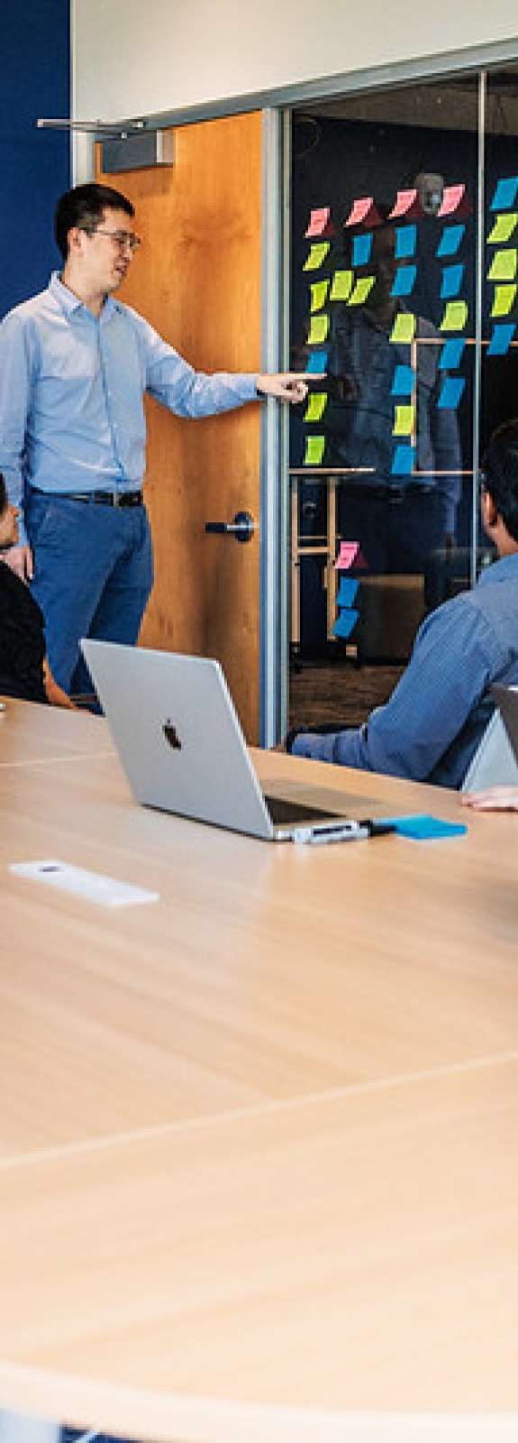 Four adults sitting at a conference table looking at another adult with sticky notes on the wall