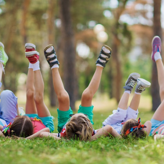 Children Lying On Green Grass In Park On A Summer Day With Their Legs Lifted Up To The Sky