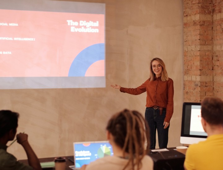 A woman in a long-sleeved shirt and pants is presenting to three students on "The Digital Evolution". The presentation is projected onto the wall, one student its in front of a PC, one has a laptop open, and one has a notebook. The room is tan with a brick wall next to it.