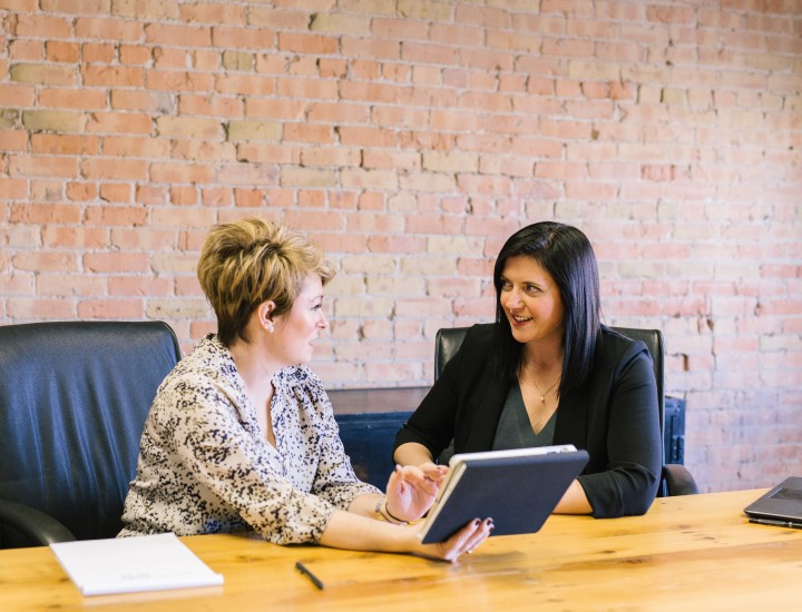 two businesswomen looking at an ipad sitting in office chairs and an office wooden meeting table with a brick wall behind them.