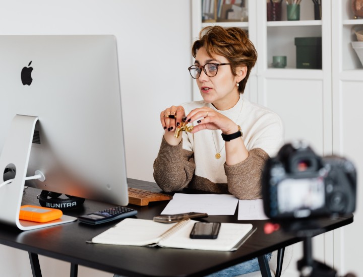 Woman sitting at a desk in front of a Apple MacBook PC. A camera is recording but out of focus in front of the desk. 